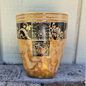 May include: A decorative ceramic vase with a textured, golden-brown finish. The upper portion features a band of black squares with gold floral and geometric designs. The rim has horizontal gold stripes. The vase is a unique home decor item.