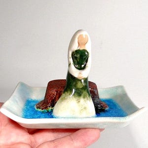May include: A ceramic figurine of a woman holding a frog, sitting on a brown log, on a white ceramic dish with blue water.