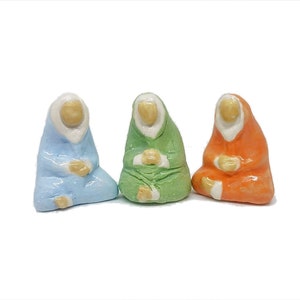 May include: Three ceramic figurines of people sitting with their hands folded in their laps. The figurines are wearing robes in different colors: light blue, green, and orange.