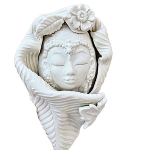 May include: A white ceramic wall hanging featuring a serene face with closed eyes, adorned with a floral crown and surrounded by stylized leaves. The sculpture has intricate details and a smooth finish.