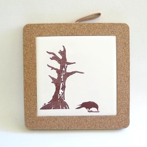 May include: A cork-framed trivet with a white ceramic tile featuring a brown silhouette of a tree and a crow. The trivet is a great addition to any kitchen.