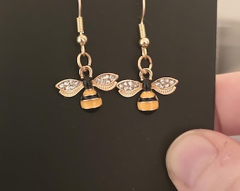 Può includere: Orecchini a forma di ape dorati con strass trasparenti. Gli orecchini sono su uno sfondo nero con il testo "HipChic" stampato sulla carta.