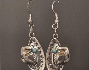 Pode incluir: Brincos de argola de prata com uma pedra turquesa e um design dourado de um chapéu de cowboy. Os brincos estão sobre um fundo preto com o texto "Hip Chic" impresso no cartão.