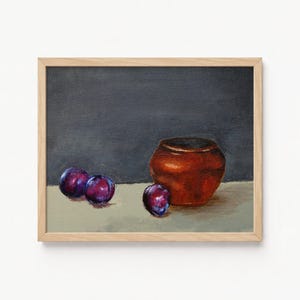 May include: Oil painting of a still life featuring a brown ceramic pot and several purple plums. The artwork is framed in a light wood frame, with a dark gray background and a light beige surface.