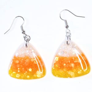 May include: A pair of Halloween-themed earrings shaped like candy corn. Each earring features a triangular design with layers of white, orange, and yellow, with glitter accents. The earrings have silver-colored fish hook ear wires.