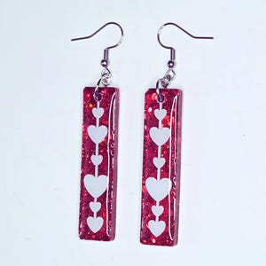 May include: A pair of red rectangular earrings with a glittery finish. Each earring features a vertical line of white hearts. The earrings have silver-colored fish hook ear wires.