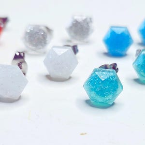 May include: A collection of colorful stud earrings with faceted, geometric designs. The earrings are in various colors, including red, pink, white, silver, blue, purple, and green. Each earring has a silver post.