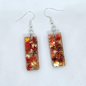 May include: A pair of silver dangle earrings with rectangular resin charms. The charms are filled with gold, red, and orange glitter and small fall leaves.