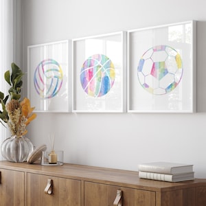 May include: Three white framed prints with watercolor illustrations of a basketball, volleyball, and soccer ball. The prints are hung on a white wall above a wooden cabinet.