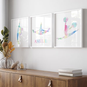 May include: Three white framed prints featuring watercolour illustrations of a cheerleader in different poses. The middle print has the name "AMELIE" written in rainbow colours below the illustration.