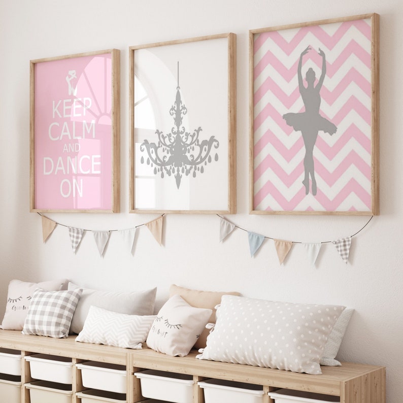 Op de afbeelding: Drie ingelijste prints op een witte muur. De linker print heeft een roze achtergrond met de tekst "Keep Calm and Dance On". De middelste print heeft een witte achtergrond met een grijs silhouet van een kroonluchter. De rechter print heeft een roze en wit chevron-patroon met een grijs silhouet van een ballerina.