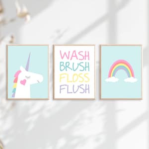 May include: Three framed prints with a pastel color scheme. The left print features a white unicorn with a rainbow mane and a pink heart on its chest. The middle print has the words "WASH BRUSH FLOSS FLUSH" in different pastel colors. The right print features a rainbow with clouds on a light blue background.