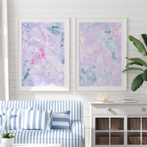 May include: Two framed abstract paintings with a purple, pink, and blue color scheme. The paintings are hanging on a white wall with white shiplap paneling.