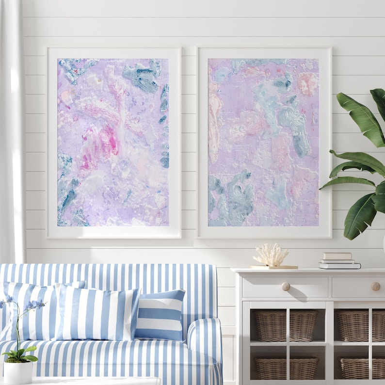 May include: Two framed abstract paintings with a purple, pink, and blue color scheme. The paintings are hanging on a white wall with white shiplap paneling.