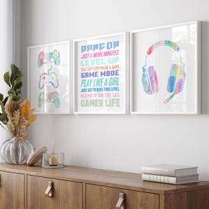 May include: A set of three framed wall art prints featuring watercolor-style gaming graphics. The left print shows three game controllers, the center print displays text including GAME ON and GAMER LIFE, and the right print shows gaming headphones.