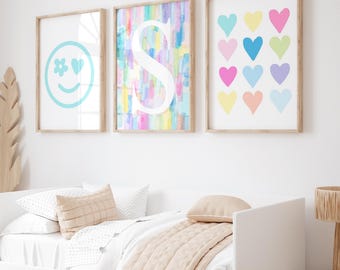 May include: Three framed prints on a white wall above a bed with a beige comforter. The prints feature a smiling face with a flower and heart, a watercolor background with the letter S, and a grid of colorful hearts.