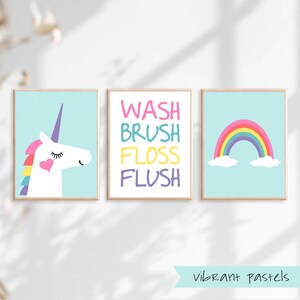 May include: Three bathroom wall art prints with a pastel color scheme. The first print features a white unicorn with a rainbow mane and a pink heart on its chest. The second print says "WASH BRUSH FLOSS FLUSH" in colorful letters. The third print features a rainbow with clouds on a light blue background. The text "vibrant pastels" is at the bottom of the image.