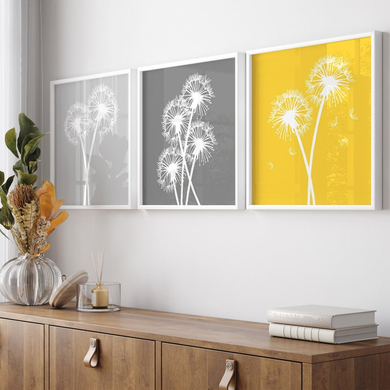 May include: Three framed prints of dandelion silhouettes on a yellow, gray, and white background. The prints are hung on a white wall above a wooden cabinet.