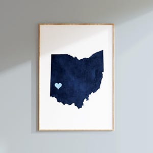 Puede incluir: Una impresión de arte mural vertical enmarcada que presenta una silueta azul marino oscuro del estado de Ohio sobre un fondo blanco liso. Un pequeño icono de corazón azul claro sólido se coloca en la parte suroeste de la forma del mapa estatal.