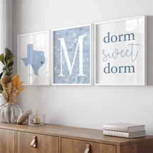 May include: Three framed wall art pieces. The left frame features a blue Texas state outline with a heart. The center frame displays a large white "M" on a blue speckled background. The right frame says "dorm sweet dorm" in blue script.