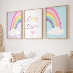 Op de afbeelding: Drie ingelijste prints met een regenboog- en wolkendesign. De middelste print heeft de naam Avery Grace in kleurrijke letters met een roze hart.
