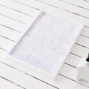 May include: A white picture frame with a light purple background. The frame is sitting on a white wooden surface.