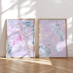 May include: Two abstract paintings in wooden frames. The paintings are both on a white background with pink, blue, and white paint splatters. The paintings are hanging on a white wall.