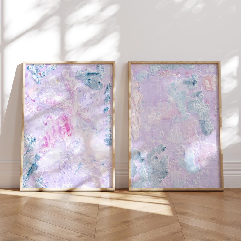May include: Two abstract paintings in wooden frames. The paintings are both on a white background with pink, blue, and white paint splatters. The paintings are hanging on a white wall.