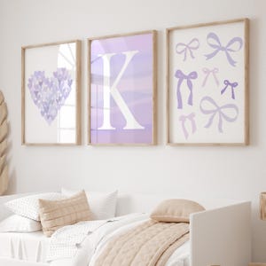 Op de afbeelding: Drie ingelijste prints met lichtbruine frames. De prints tonen een hart gemaakt van paarse harten, een witte letter 'K' op een paarse achtergrond en diverse paarse strikken. Het kunstwerk bevindt zich boven een bed met kussens en een deken.
