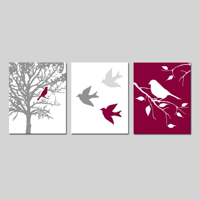 Puede incluir: Conjunto de tres impresiones de arte de pared con un &aacute;rbol gris con un p&aacute;jaro rojo, tres p&aacute;jaros grises en vuelo y un p&aacute;jaro blanco posado en una rama sobre un fondo burdeos.