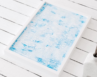 Op de afbeelding: Een wit frame met een blauw en wit abstract schilderij erin. Het frame staat op een wit houten oppervlak.