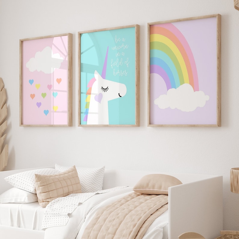 May include: Three framed art prints with pastel backgrounds. One features a cloud and hearts, another a unicorn with text, and the last a rainbow. The prints are in wooden frames and hang above a bed with neutral-toned bedding.
