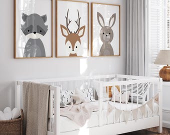 May include: Three framed animal art prints featuring a raccoon, deer, and rabbit. The illustrations are in neutral tones of gray, brown, and white, with wooden frames. The prints are displayed above a white crib with a beige blanket and decorative pillows.