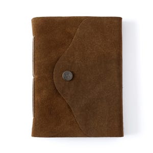 Op de afbeelding: Een bruin suède notitieboek met een klep sluiting en een metalen knoop. Het notitieboek heeft een rustieke en vintage uitstraling.