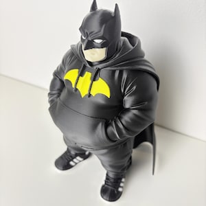 May include: A 3D printed figurine of a character wearing a black hooded sweatshirt with a yellow bat emblem on the chest, black trousers, and black trainers with white stripes. The figure has a black cowl with pointed ears and white eye slits.