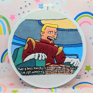 Könnte beinhalten: Stickbild in einem Stickrahmen mit einem Zeichentrickcharakter aus Futurama, Zapp Brannigan, in einer rot-goldenen Uniform. Der Text unter ihm lautet "Das ist das Gesetz, Leela, und Brannigans Gesetz ist wie Brannigans Liebe - hart und schnell."