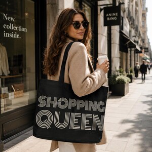 Könnte beinhalten: Schwarze Tragetasche mit dem weißen Schriftzug "SHOPPING QUEEN". Eine Person trägt die Tasche, bekleidet mit einem beigen Mantel und Sonnenbrille. Die Tasche hat ein schlichtes, auffälliges Design, ideal zum Einkaufen.