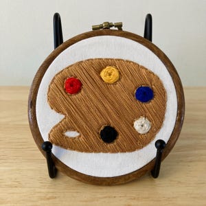 May include: A brown wooden embroidery hoop with a white fabric background. The fabric has an embroidered image of a brown paint palette with colorful paint dots in red, yellow, blue, white, and black.