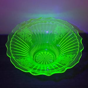 May include: A vibrant, glowing neon green glass bowl featuring a scalloped rim and a radiating ribbed pattern. The interior surface is decorated with etched floral motifs, and the base displays a starburst design, all illuminated under UV light.