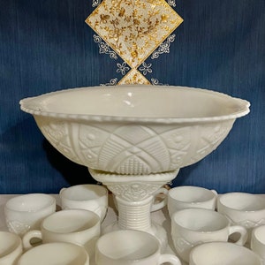 May include: A white milk glass punch bowl with a pedestal base and a decorative embossed pattern. The bowl is surrounded by 10 smaller white milk glass cups.