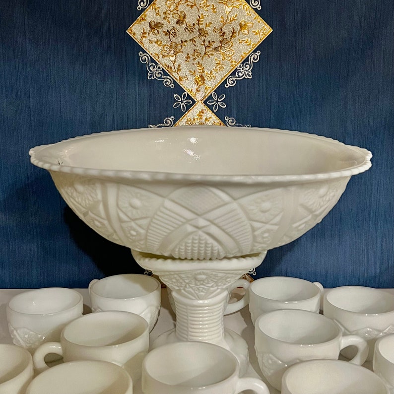 May include: A white milk glass punch bowl with a pedestal base and a decorative embossed pattern. The bowl is surrounded by 10 smaller white milk glass cups.