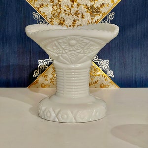 May include: A white milk glass pedestal bowl with a scalloped edge and a floral design. The bowl is on a pedestal base with a fluted design.