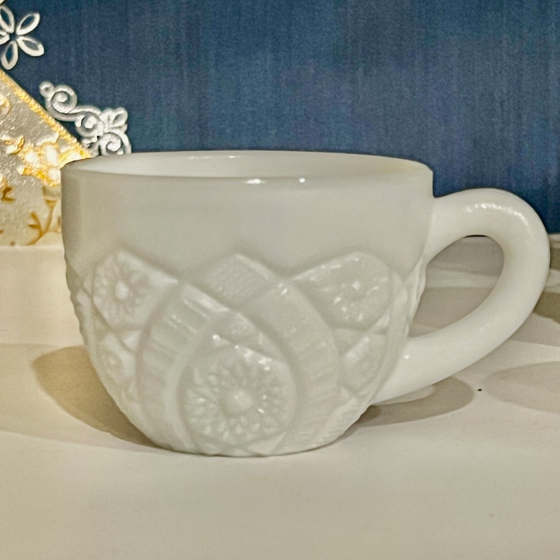 May include: A white milk glass cup with a raised floral design. The cup has a handle and is perfect for serving beverages.