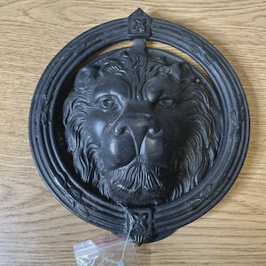 May include: A circular black metal door knocker featuring a detailed, three-dimensional lion head sculpture centered within a decorative ring frame. The lion has a prominent mane and facial features, and the frame includes small floral accents.