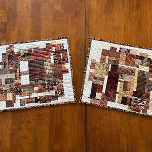 May include: Two rectangular quilted placemats with a patchwork design. The placemats feature a variety of brown, beige, and cream fabrics arranged in a geometric pattern. Each placemat has a white border and a patterned brown edge.