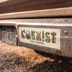 May include: A rectangular bumper sticker with a cream background featuring the word COEXIST in large, stylized letters filled with nature scenes including mountains, a bear, a river, a deer, an owl, trees, and flowers, with the text with wildlife.