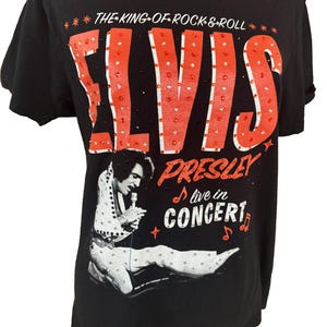 May include: A black short-sleeved t-shirt featuring a graphic of Elvis Presley in a white jumpsuit holding a microphone. The text reads THE KING OF ROCK & ROLL, ELVIS, PRESLEY, and live in CONCERT. The design is accented with small red rhinestones.