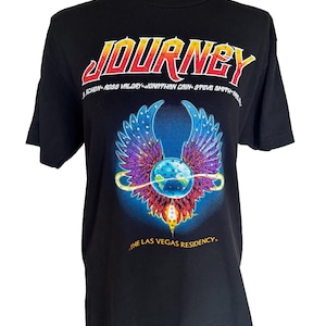 以下が含まれることがあります： バンドJourneyのグラフィックがプリントされた黒の半袖Tシャツ。赤と黄色の「JOURNEY」の文字、青と紫のアクセントが入った翼のある地球儀のエンブレム、そして「THE LAS VEGAS RESIDENCY」というテキストがデザインされています。