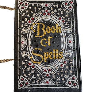 May include: Black Book of Spells handbag with gold chain strap. The cover features the words "Book of Spells" in gold lettering, surrounded by white Celtic knot designs, red and green gems, and small silver embellishments. A unique accessory.