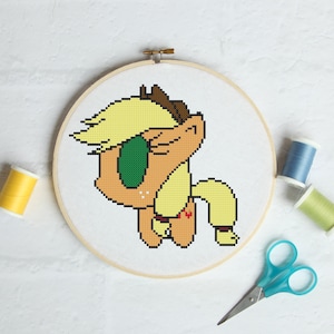 Puede incluir: Patrón de punto de cruz de un pony de dibujos animados con pelo amarillo, un ojo verde y una manzana roja en el costado. El pony está en un aro sobre un fondo blanco.